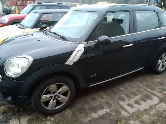 Czarny samochód Mini Cooper SUV z widocznym napisem 'ALL4' na boku, zaparkowany na betonowej powierzchni z plamami. W tle widoczne są inne pojazdy: żółty i czerwony samochód. Samochód ma srebrne felgi, czarne zderzaki i lusterka. Tło obejmuje fragment terenu z trawą oraz budynki w oddali.