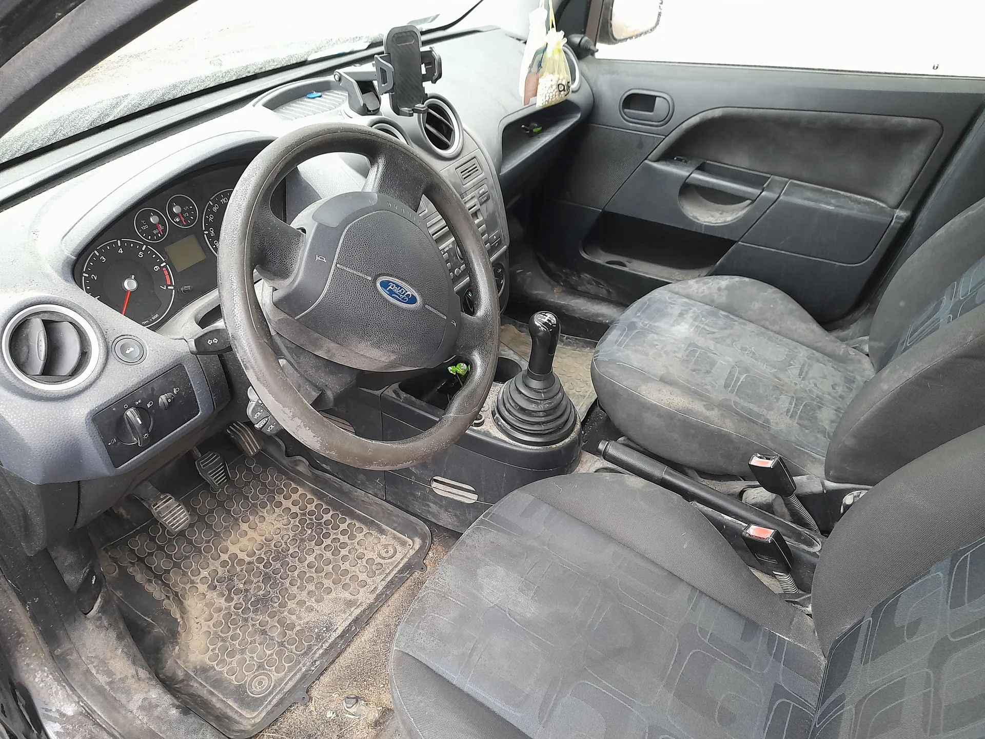 Wnętrze samochodu marki Ford z widocznym kierownicą z logo, tablicą przyrządów z licznikami prędkości i obrotomierzem, dźwignią zmiany biegów, czarnymi siedzeniami z geometrycznym wzorem, brudnymi dywanikami podłogowymi oraz uchwytem na telefon na desce rozdzielczej. W tle widoczna drzwi pasażerskie i woreczek wiszący na lusterku wstecznym.