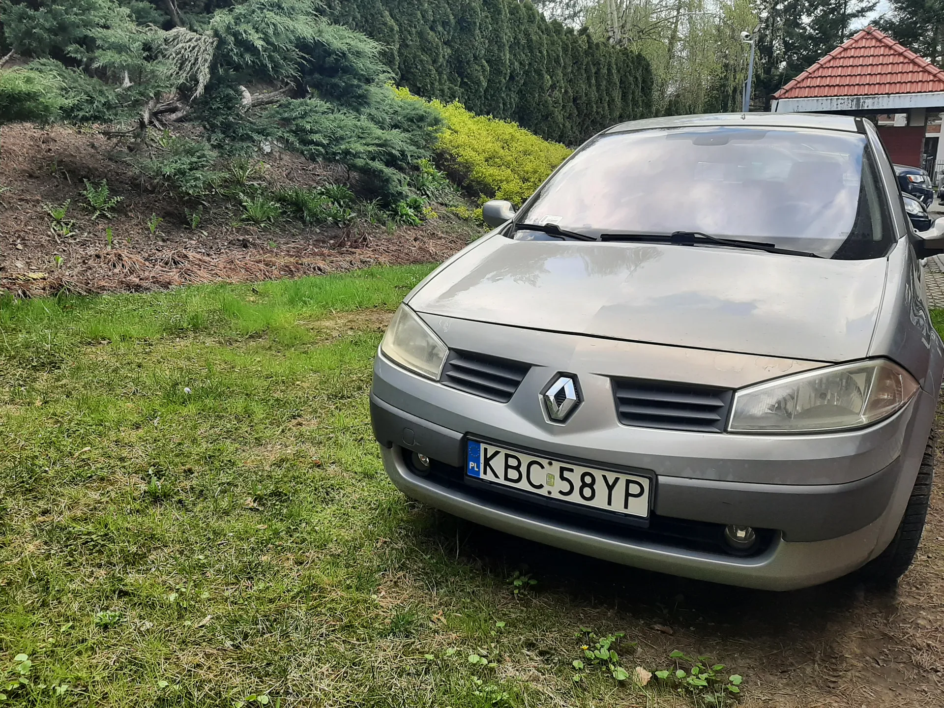 Srebrny samochód marki Renault zaparkowany na trawie. Widoczna tablica rejestracyjna z polskim kodem PL i numerem KBC 58YP. W tle zielone krzewy, drzewa oraz budynek z czerwoną dachówką. Samochód znajduje się na trawiastym obszarze z widocznymi śladami ziemi i roślinności.