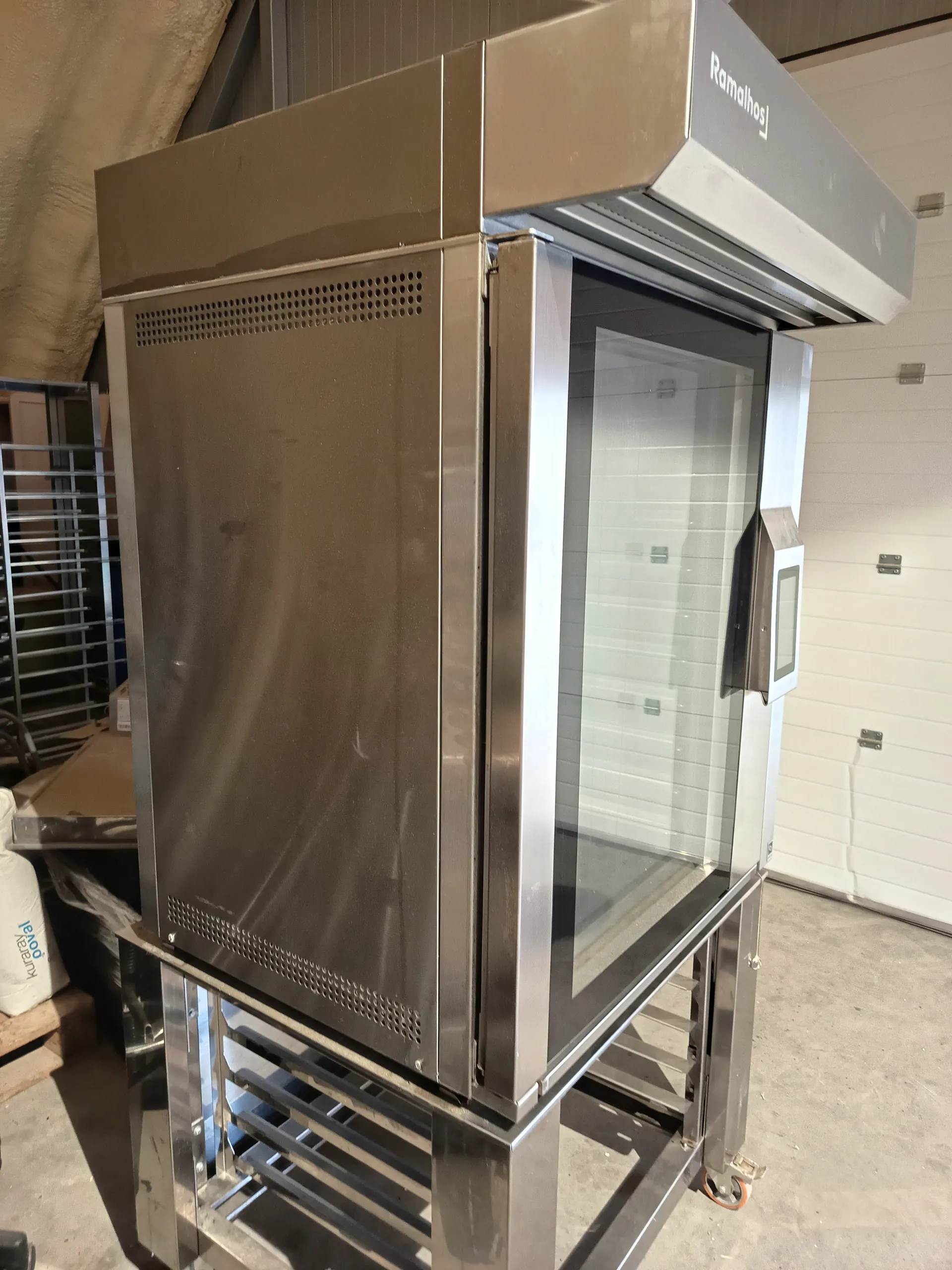 Przemysłowy, stalowy aparat z szklanymi drzwiami i panelem sterowania. Na górze widoczny napis 'Ramalhos'. W tle biała ściana, metalowa półka oraz biała torba z napisem 'Knauf'. Podłoga betonowa.
