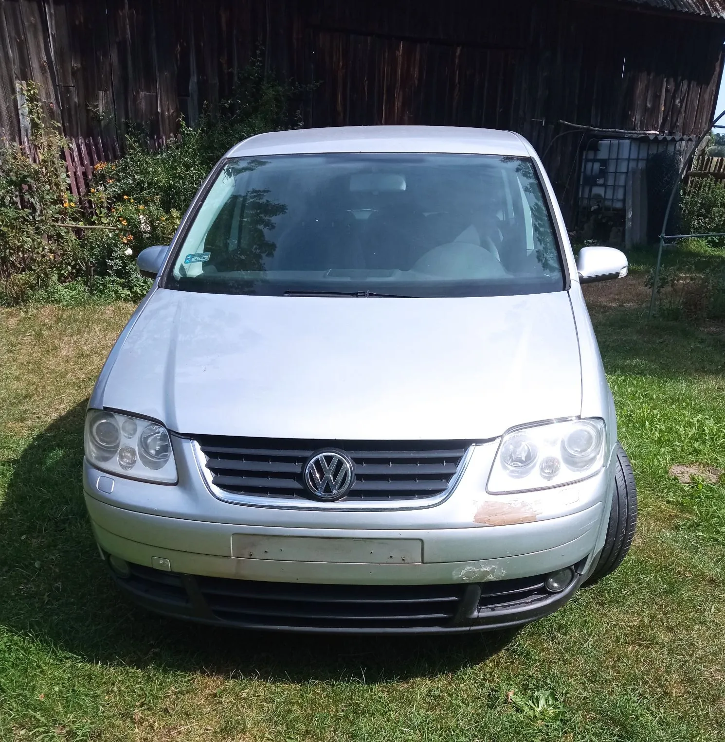 Srebrny samochód marki Volkswagen widziany od przodu, zaparkowany na trawie na tle drewnianego budynku.