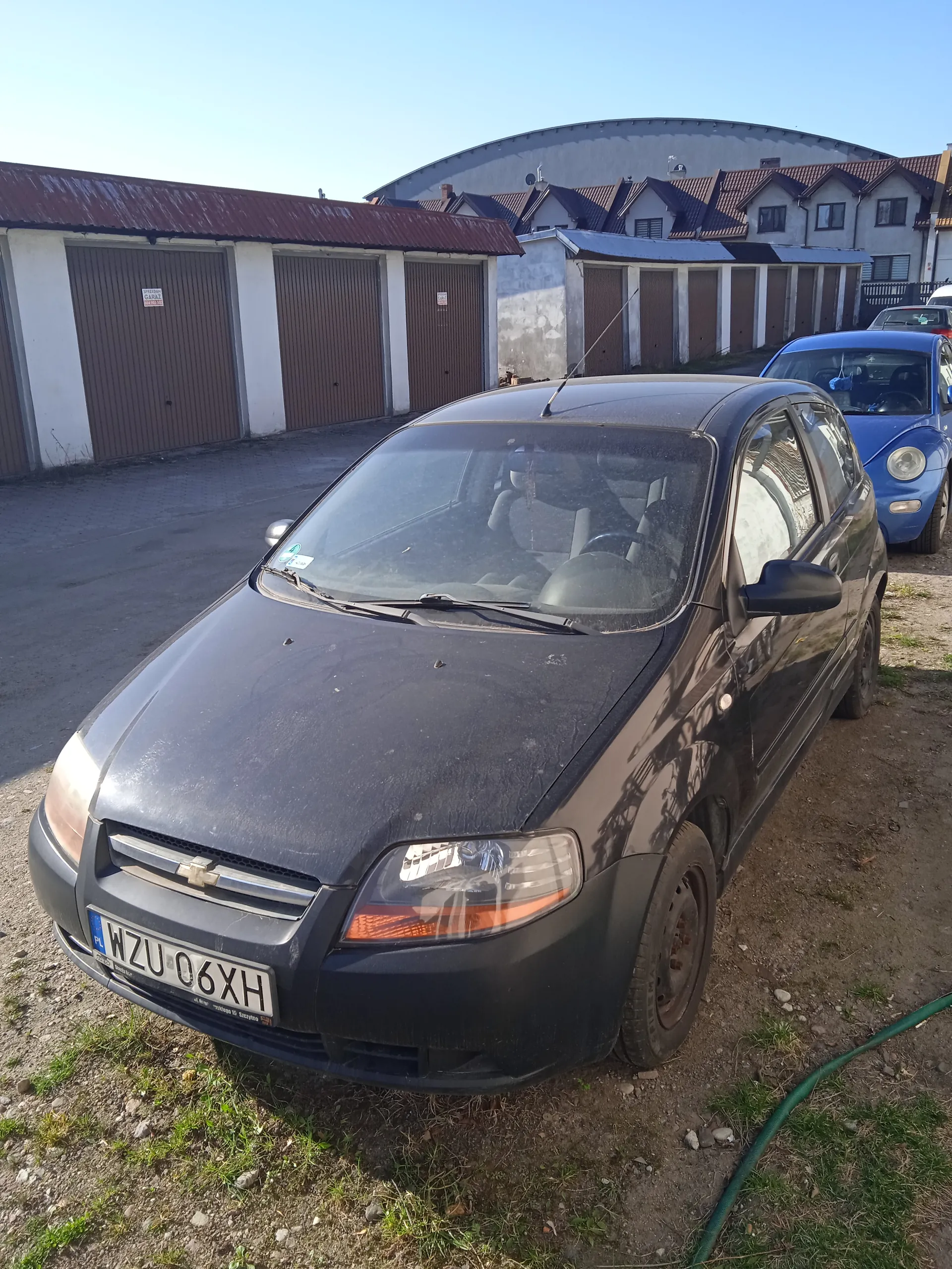 Czarny samochód marki Chevrolet z tablicą rejestracyjną WZU 06XH zaparkowany na podwórku z drobnej ziemi i trawy. W tle widoczne są garaże z brązowymi drzwiami, budynek z zakrzywionym dachem oraz niebieski samochód. Na niebie nie ma chmur.