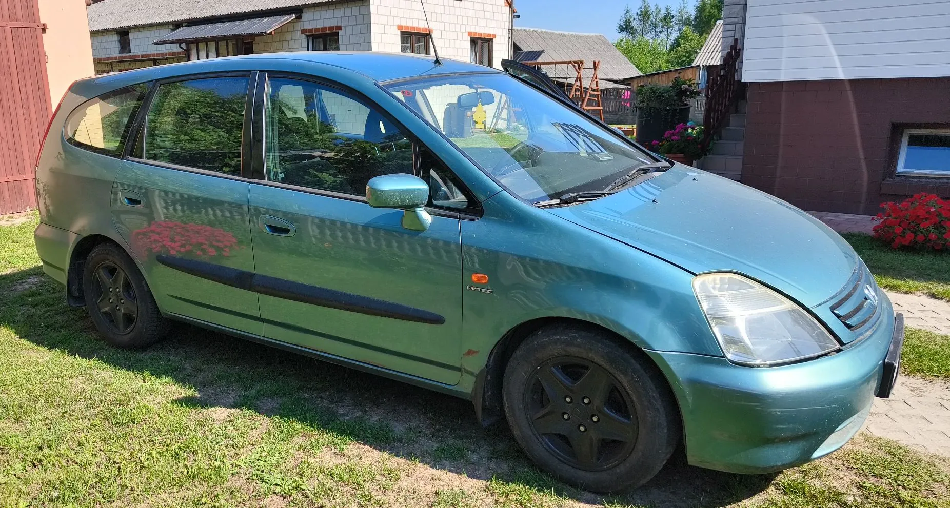 Zielony samochód typu kombi zaparkowany na trawie przed domami. Pojazd ma czarne felgi i czarne listwy ochronne na drzwiach. Na boku samochodu widoczny jest napis 'i-VTEC'. W tle widać fragmenty budynków oraz roślinność.