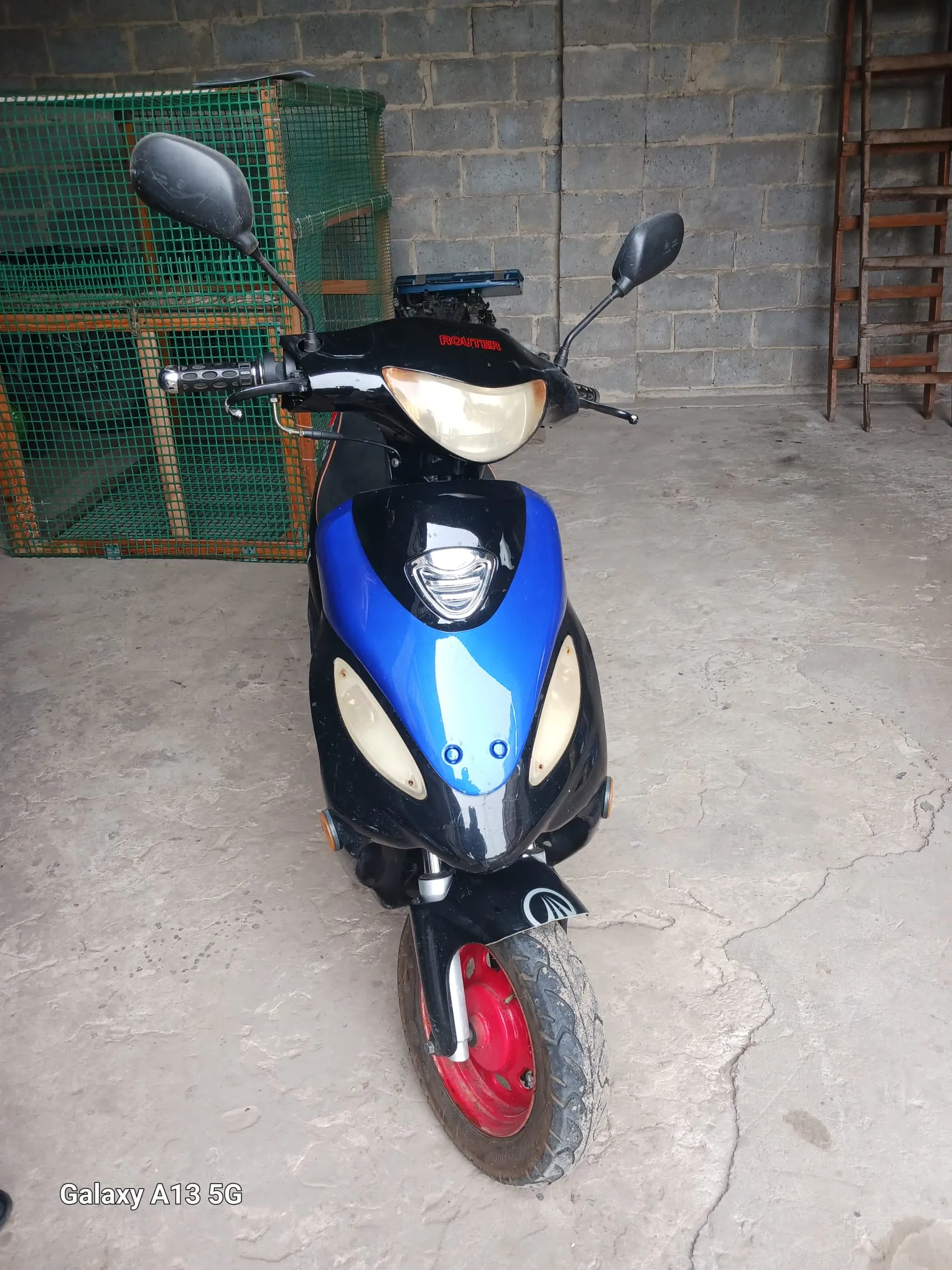 Scooter o kolorze czarno-niebieskim z czerwonymi felgami, widoczny z przodu. Na masce napis 'ROUTER' czerwonymi literami. Po bokach lusterka wsteczne, reflektor główny i dwa boczne światła. Tło: szara betonowa ściana, zielona siatka oraz drewniana drabina. Na dole zdjęcia widoczny napis 'Galaxy A13 5G'. Podłoga z betonu z widocznymi pęknięciami.
