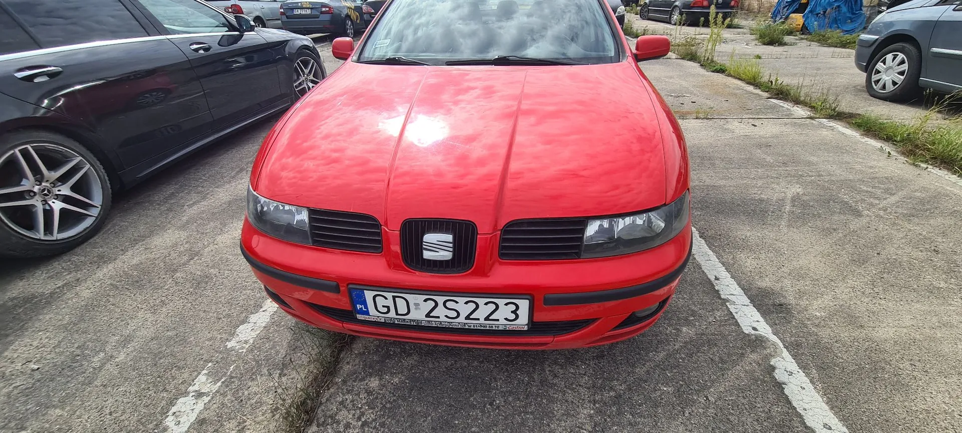 Widok z przodu czerwonego samochodu marki Seat zaparkowanego na betonowym parkingu. Samochód ma polską tablicę rejestracyjną o numerze GD 2S223. Po lewej stronie widoczny jest fragment czarnego samochodu, a w tle inne pojazdy i pas zieleni.