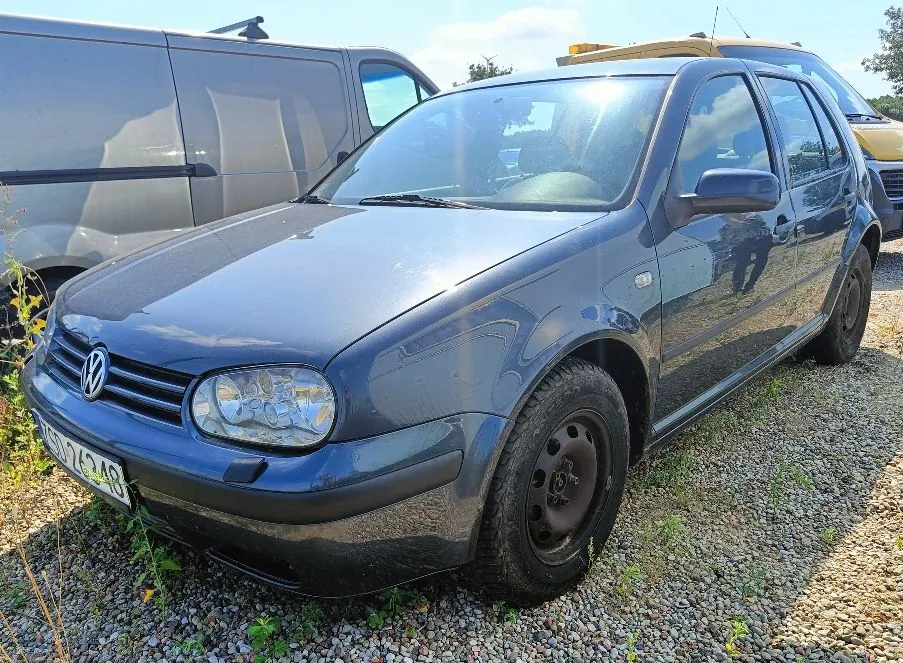 Srebrno-niebieski samochód osobowy marki Volkswagen Golf zaparkowany na żwirowym podłożu. Widoczny jest przód i prawy bok auta. W tle znajduje się srebrny furgon. Na przednim zderzaku widoczna jest fragment tablicy rejestracyjnej z numerem kończącym się na 248.