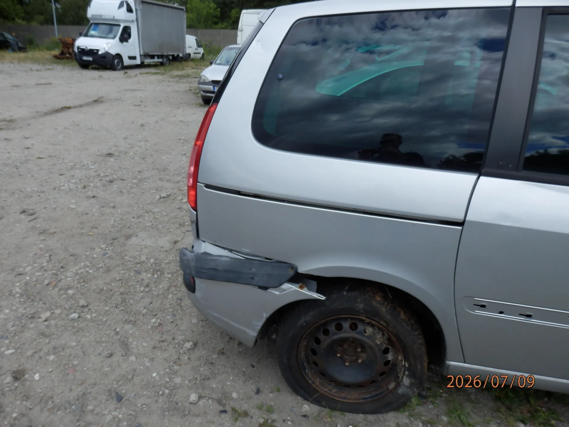 Zdjęcie przedstawiające tylną, lewą część srebrnego samochodu osobowego z widocznymi uszkodzeniami. Zderzak jest wgnieciony i częściowo oddzielony od karoserii, a nad nadkolem znajduje się wklęsłe wgniecenie. W tle widoczny jest żwirowy plac, biały samochód osobowy oraz biała ciężarówka z zabudową kontenerową. W prawym dolnym rogu znajduje się pomarańczowy napis z datą: 2026/07/09.