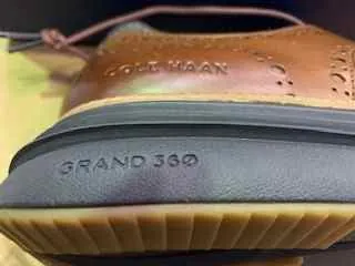 Zbliżenie brązowego skórzanej buta z widocznym wygrawerowanym napisem "COLE HAAN" na górnej części. Na pięcie umieszczony jest napis "GRAND 360" na ciemnej warstwie. Podeszwa posiada warstwową konstrukcję w kolorach czarnym i beżowym z teksturą. Tło stanowi rozmyta zielonkawa powierzchnia. But ma widoczne przeszycia i oczka.