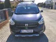 Zdjęcie przodu szarego samochodu osobowego marki Dacia, zaparkowanego na kostce brukowej. Pojazd posiada zamontowany bagażnik dachowy oraz tablicę rejestracyjną o treści PL 7805E.