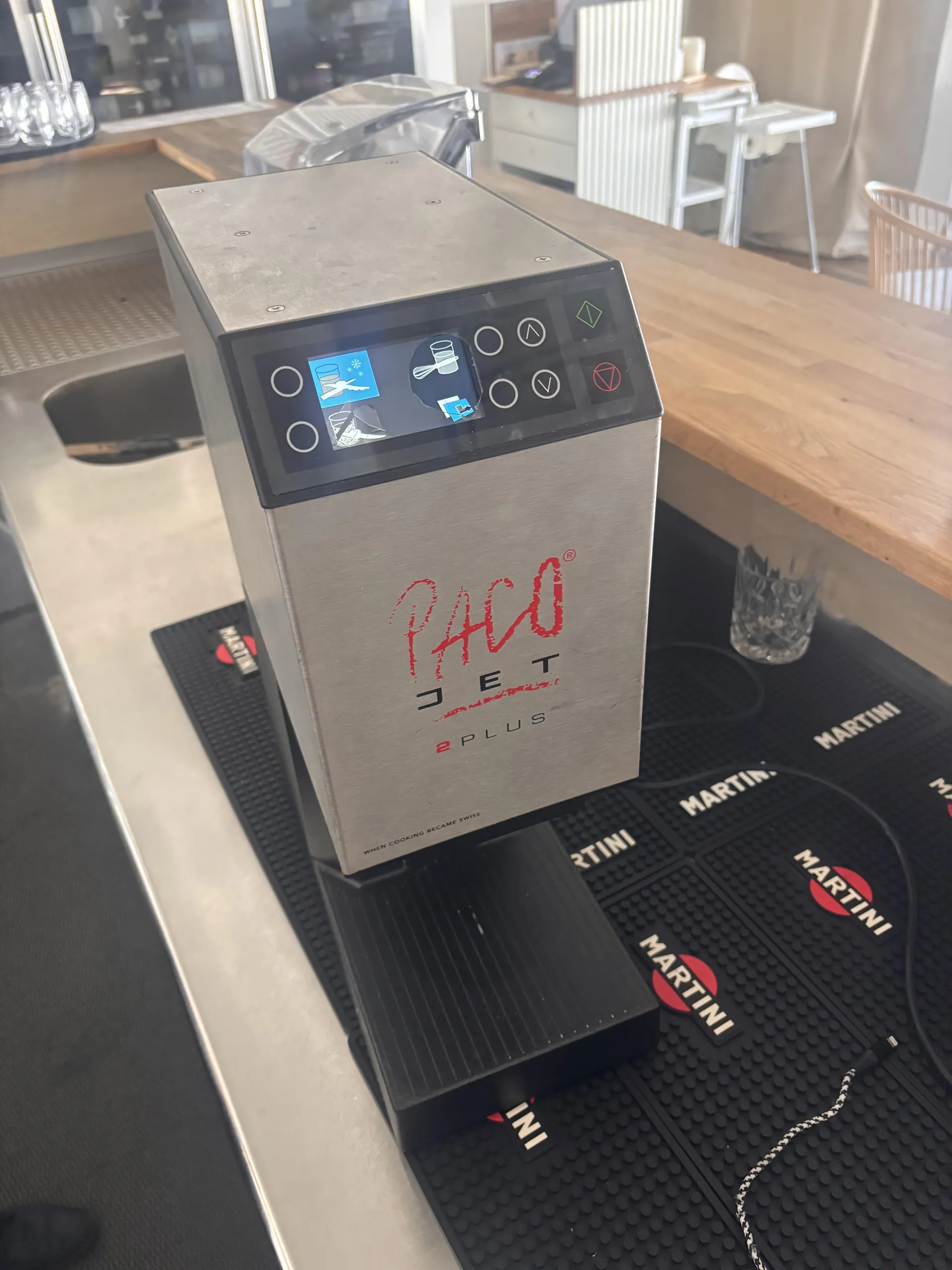 Srebrny automat z ekranem dotykowym i przyciskami, na którym widnieje czerwony napis 'PACO JET 2 PLUS'. Automat stoi na czarnej podkładce z powtarzającym się logo 'MARTINI' w białym i czerwonym kolorze. W tle widoczny jest drewniany blat baru, zlew, szklanki oraz krzesła. Na ekranie automatu znajdują się ikony, w tym niebieska ikona z lodem i szklanką.