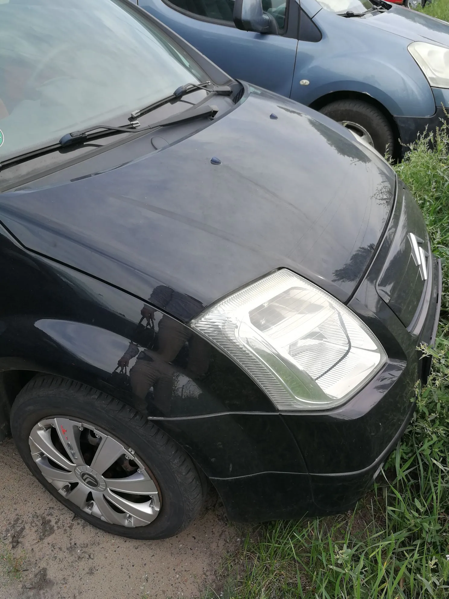 Zbliżenie na przód czarnego samochodu osobowego, widoczna jest maska, prawa lampa przednia oraz przednie prawe koło z felgą aluminiową z logo Citroën. W tle widoczny jest fragment innego, niebieskiego samochodu oraz zielona trawa.
