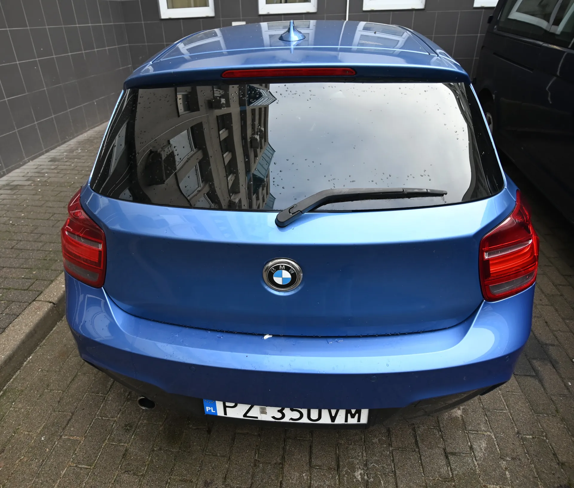 Niebieski samochód BMW z tyłu, tablica rejestracyjna PL PZ 35UVM. Mokre tylne szyby z kroplami deszczu i wycieraczką, czerwone światła, logo BMW, antena dachowa. Tło: szara ściana z białymi oknami, brukowana droga, czarny samochód po prawej.