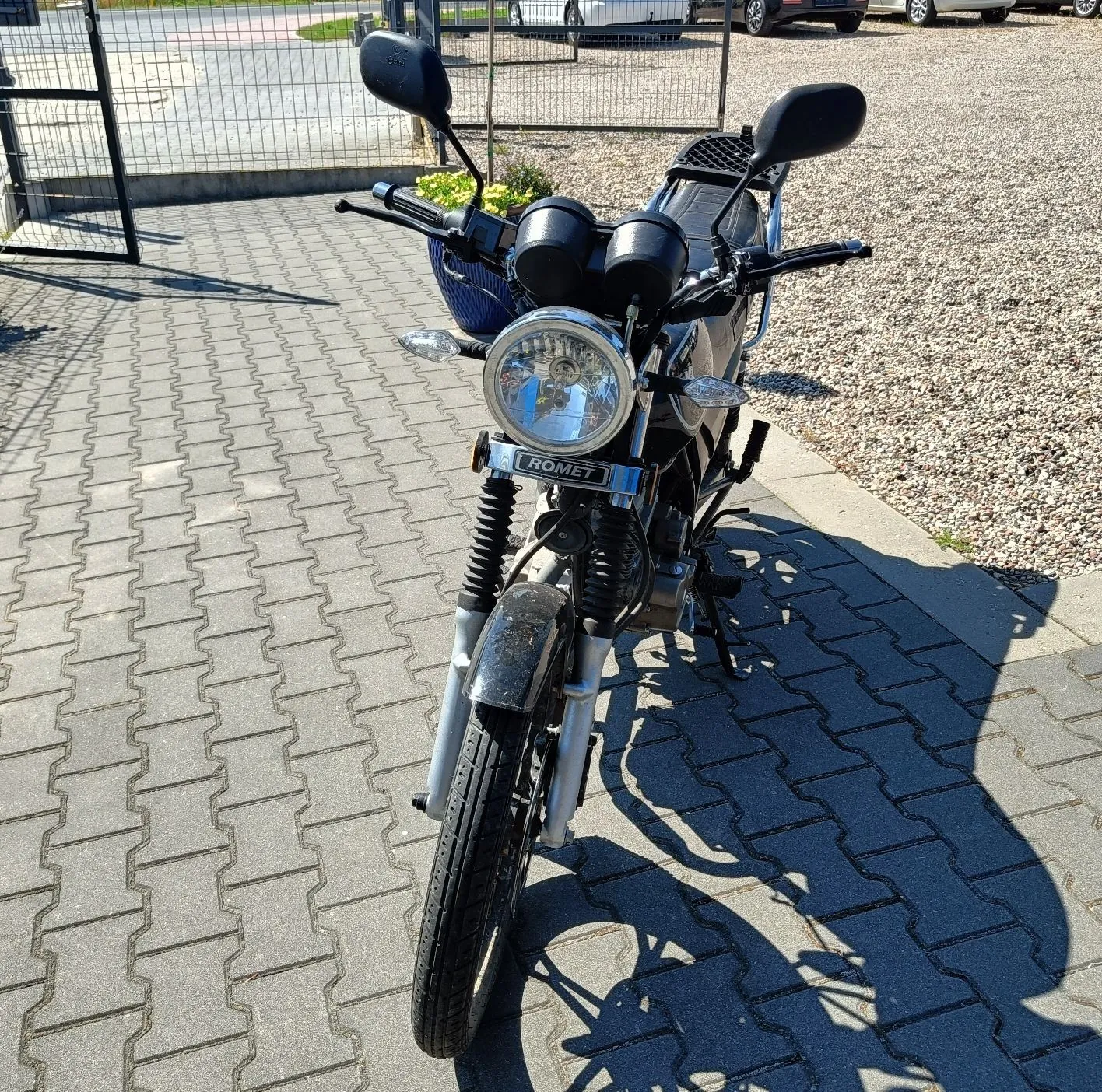 Widok z przodu na czarny motocykl marki Romet, zaparkowany na szarym kostrukowym podjeździe. Motocykl posiada okrągły reflektor z napisem ROMET na błotniku pod lampą, dwa lusterka wsteczne oraz czarną kierownicę. W tle widoczny jest metalowy płot oraz fragmenty białych samochodów na żwirowym parkingu.
