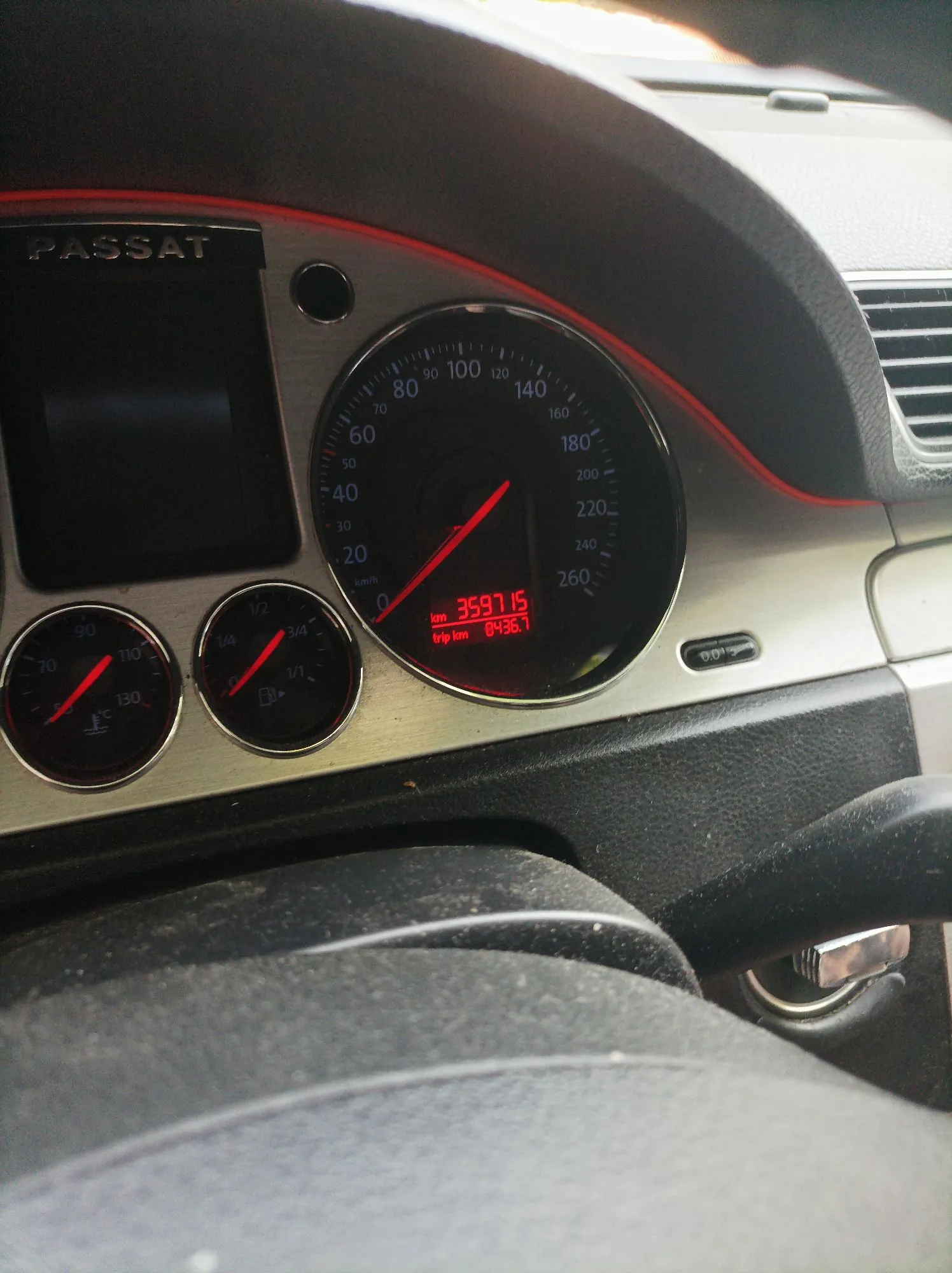 Zbliżenie na deskę rozdzielczą samochodu marki Passat. Widoczny jest okrągły prędkościomierz z cyframi od 0 do 260 km/h, z czerwoną wskazówką. W dolnej części prędkościomierza znajduje się cyfrowy wyświetlacz z pomarańczowym tekstem: 'km 35971.5' oraz 'trip km 0436.7'. Po lewej stronie znajdują się dwa mniejsze, okrągłe wskaźniki: obrotomierz oraz wskaźnik temperatury cieczy chłodzącej. Nad prędkościomierzem widnieje napis 'PASSAT'.