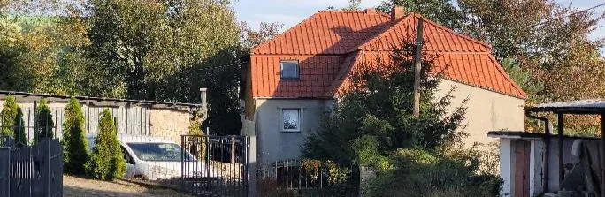 Dom z czerwoną dachówką, biały samochód zaparkowany przy drewnianym płocie, zielone krzewy i drzewa z jesiennymi liśćmi, kamienna ściana po lewej stronie, metalowa brama, niewielka budowla po prawej stronie, jasne niebo.