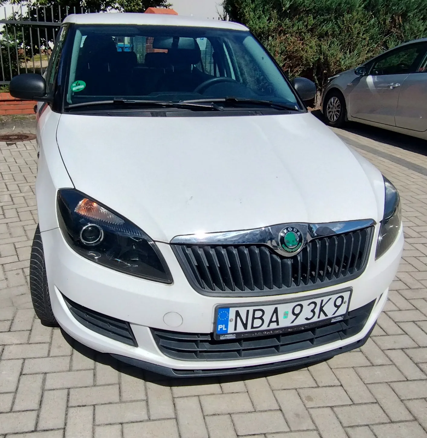 Biały samochód marki Skoda zaparkowany na kostce brukowej. Widoczny jest przód pojazdu z polską tablicą rejestracyjną o numerze NBA 93K9. W tle widać zielone krzewy oraz fragment innego, srebrnego samochodu.