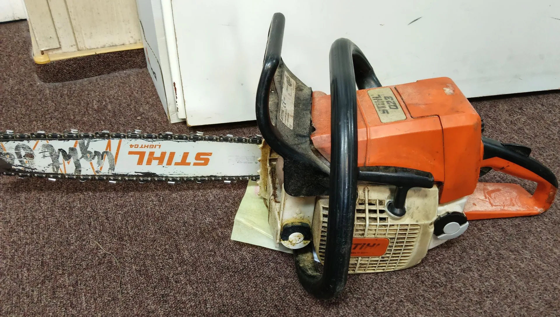 Pomarańczowa piła spalinowa marki STIHL leżąca na brązowej wykładzinie. Urządzenie posiada długą prowadnicę z łańcuchem, na której widnieje biały napis 'STIHL' oraz mniejszy napis 'LIGHT 04'. Piła ma czarny uchwyt i białą obudowę silnika z logo marki.