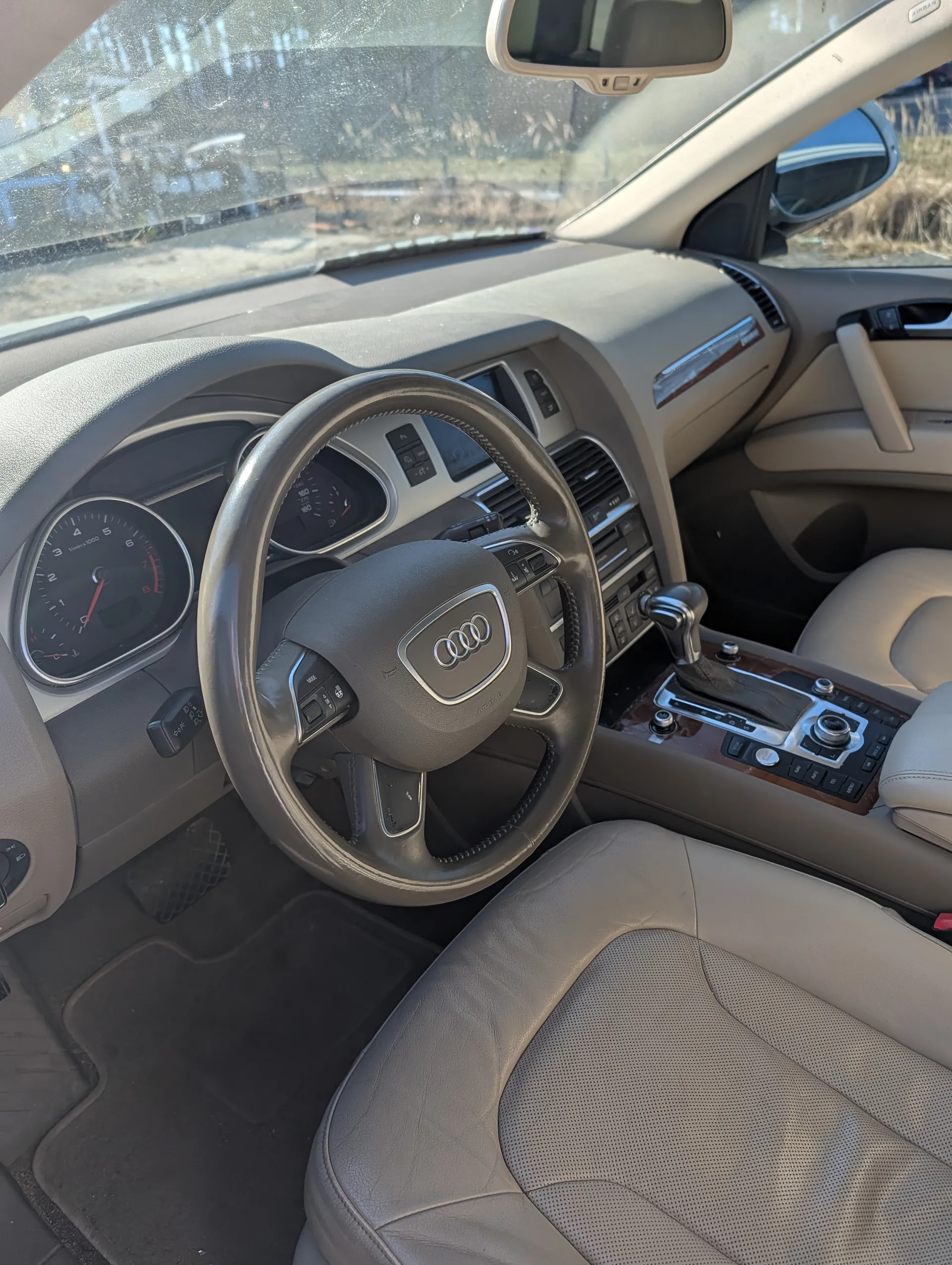 Wnętrze samochodu marki Audi z widocznym kierownicą z logo Audi, tablicą przyrządów z licznikiem obrotów i prędkościomierzem, beżowymi skórzonymi fotelami, centralną konsolą z przyciskami i dźwignią zmiany biegów. Przez przednią szybę widoczny zewnętrzny krajobraz z suchą trawą i drogą.