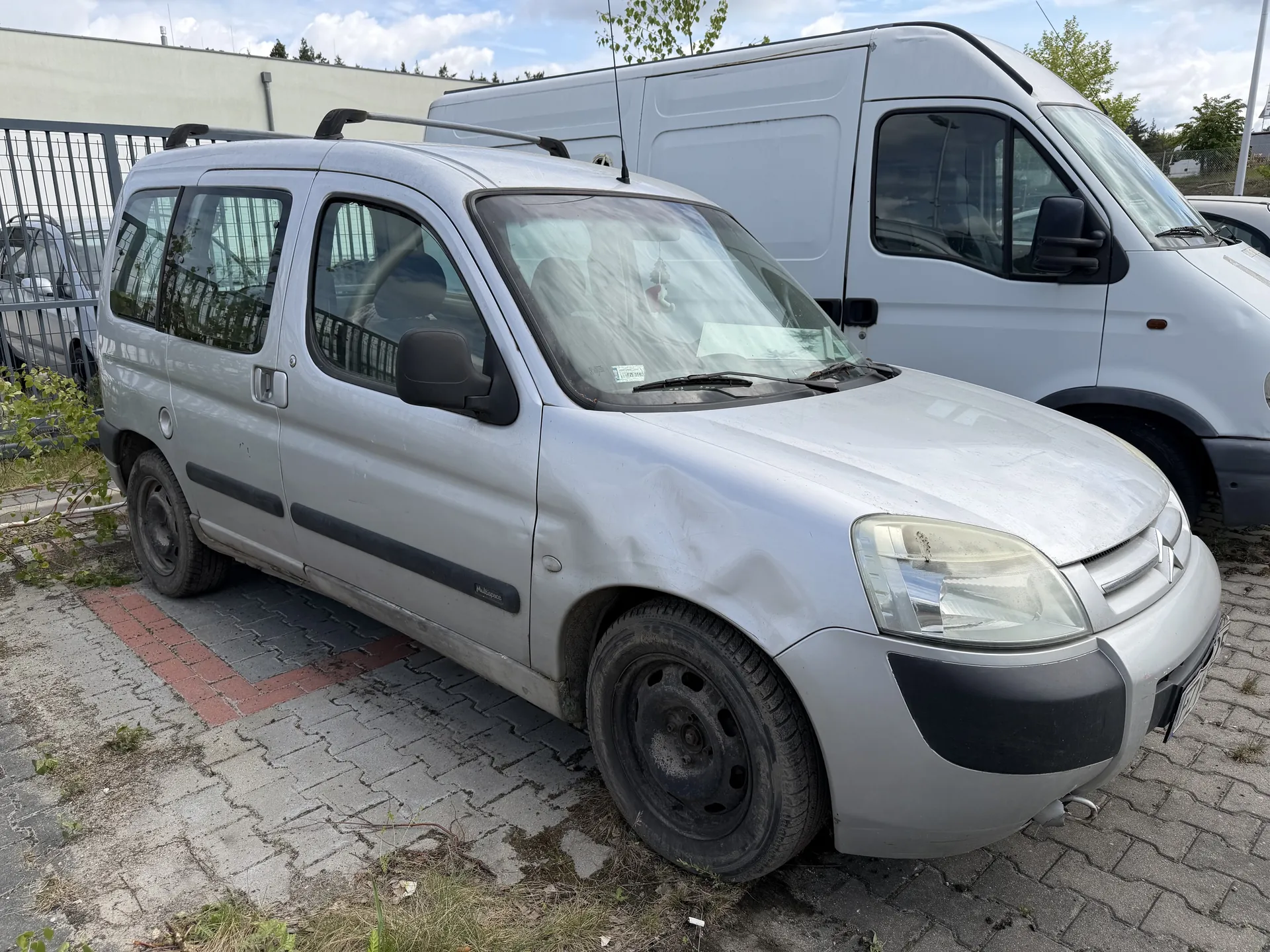 Srebrny samochód dostawczy z czarną listwą boczną i dachowym bagażnikiem, z widocznym wgnieceniem na przedniej prawej stronie. Pojazd stoi na brukowanym placu z czerwonymi i szarymi płytami, obok metalowego ogrodzenia. W tle biały samochód dostawczy, budynek o jasnej elewacji oraz drzewa. Niebo częściowo zachmurzone. Na przedniej szybie srebrnego pojazdu widoczny mały naklejony dokument.