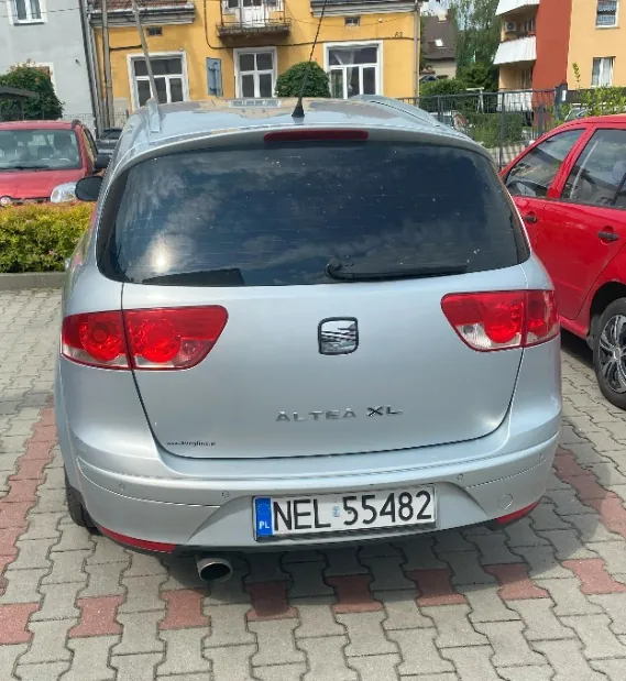 Srebrny samochód marki Seat Altea XL zaparkowany tyłem na kostce brukowej. Na klapie bagażnika widoczne jest logo marki oraz napis 'ALTEA XL'. Samochód posiada polską tablicę rejestracyjną o numerze NEL 55482. W tle widać czerwony samochód po prawej stronie, fragment czerwonego auta po lewej oraz żółty budynek mieszkalny.