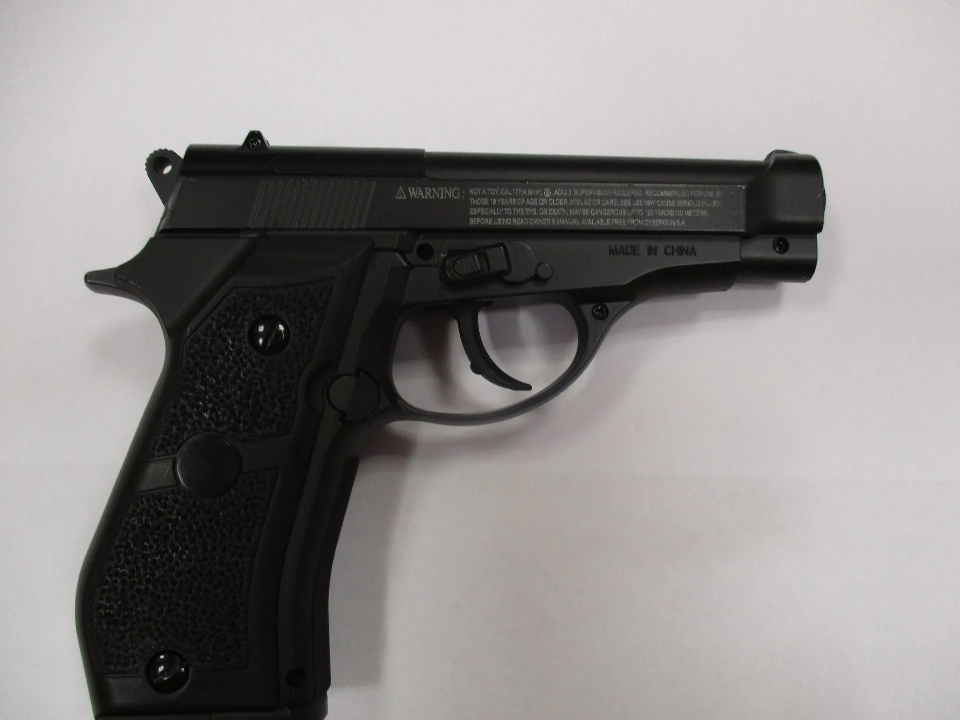 Czarny pistolet wykonany z tworzywa sztucznego, przedstawiony z profilu na jasnym, neutralnym tle. Na górnej części zamka znajduje się napis: 'WARNING: NOT A TOY. ONLY FOR 6mm BBs. ADULT SUPERVISION IS REQUIRED. RECOMMENDED FOR USE BY PERSONS 18 YEARS OF AGE OR OLDER. MISUSE OR CARELESS USE MAY CAUSE SERIOUS INJURY, ESPECIALLY TO THE EYE, OR DEATH. MAY BE DANGEROUS UP TO 20m/60ft (10m/30m METERS). BEFORE USING READ OWNER'S MANUAL. AVAILABLE FREE FROM CYBORGUN S.A.'. Poniżej, po prawej stronie, widnieje napis 'MADE IN CHINA'. Rękojeść pistoletu ma chropowatą teksturę.