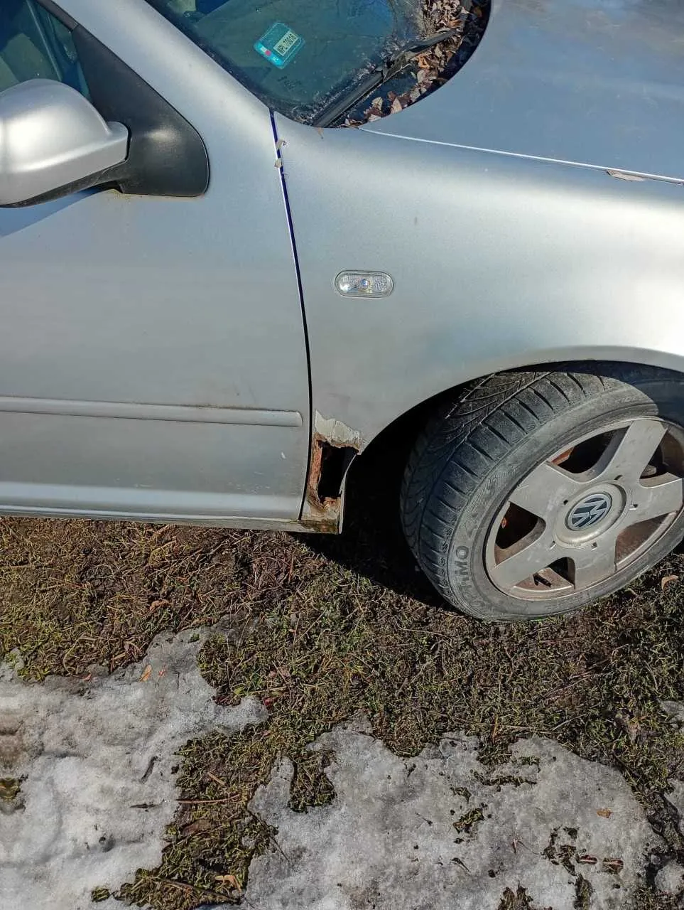 Srebrny samochód marki Volkswagen z widocznym uszkodzeniem i rdzą w okolicy tylnego błotnika. Na przednim szybie widoczny jest niebieski znaczek rejestracyjny. Samochód stoi na trawie z resztkami śniegu. Widoczne są lusterko boczne, reflektor na drzwiach oraz felga z logo VW.