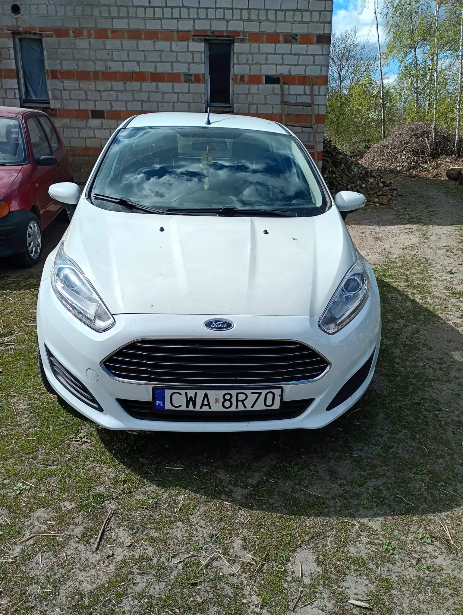 Biały samochód marki Ford z tablicą rejestracyjną CWA 8R70 (PL) na pierwszym planie. Po lewej stronie częściowo widoczny czerwony samochód. W tle budynek z cegły i bloczków z niezakonczonymi oknami, stos drewna i drzewa. Niebo częściowo zachmurzone. Na tablicy rejestracyjnej widoczny napis CWA 8R70 oraz flaga Polski (PL).