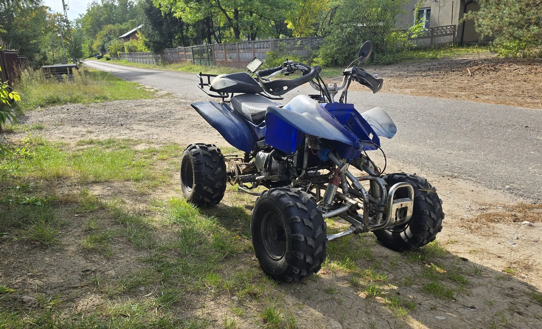 Niebieski quad zaparkowany na poboczu szutrowej drogi. Pojazd ma duże, terenowe opony i jest ustawiony przodem w prawą stronę obrazu. W tle widoczna jest droga prowadząca w głąb, zielone drzewa, krzewy oraz fragment ogrodzenia i budynku.