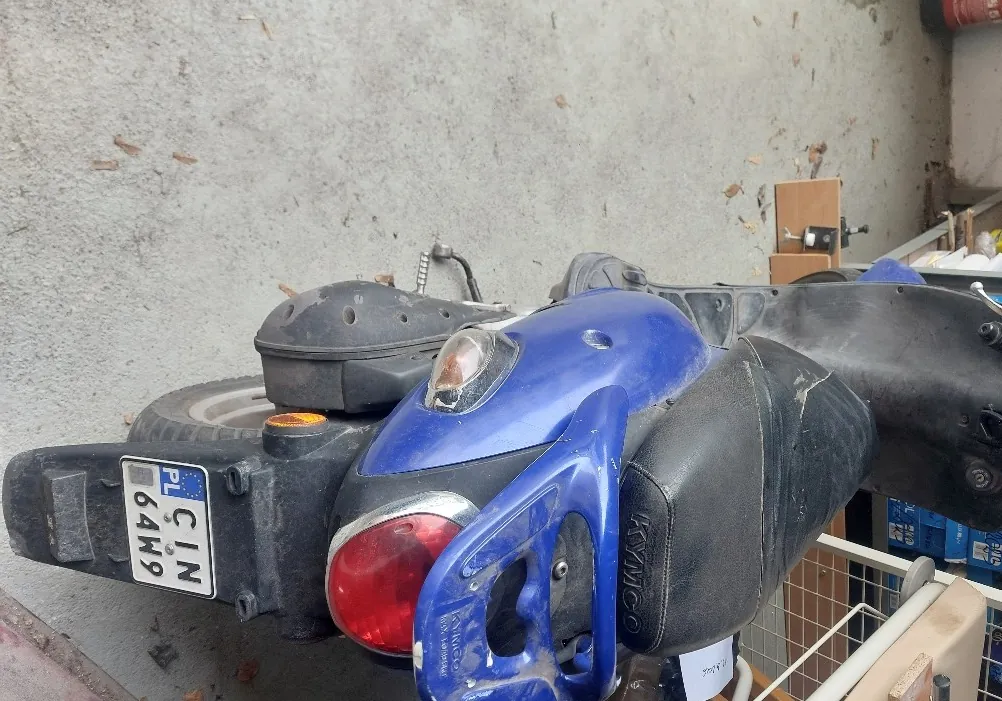 Zdjęcie przedstawia niebieski skuter marki KYMCO z widocznym tyłem. Na czarnym siedzeniu widnieje napis "KYMCO". W dolnej części obrazu znajduje się tablica rejestracyjna z białym tłem i czarnym tekstem: "PL CIN 64W9". Skuter posiada czerwone światło tylnego oświetlenia oraz pomarańczowe kierunkowskazy. Tło stanowi betonowa podłoga z rozrzuconymi liśćmi oraz fragmenty pomieszczenia z półkami i przedmiotami w tle. Siedzenie skutera jest zużyte, z widocznymi śladami zużycia.