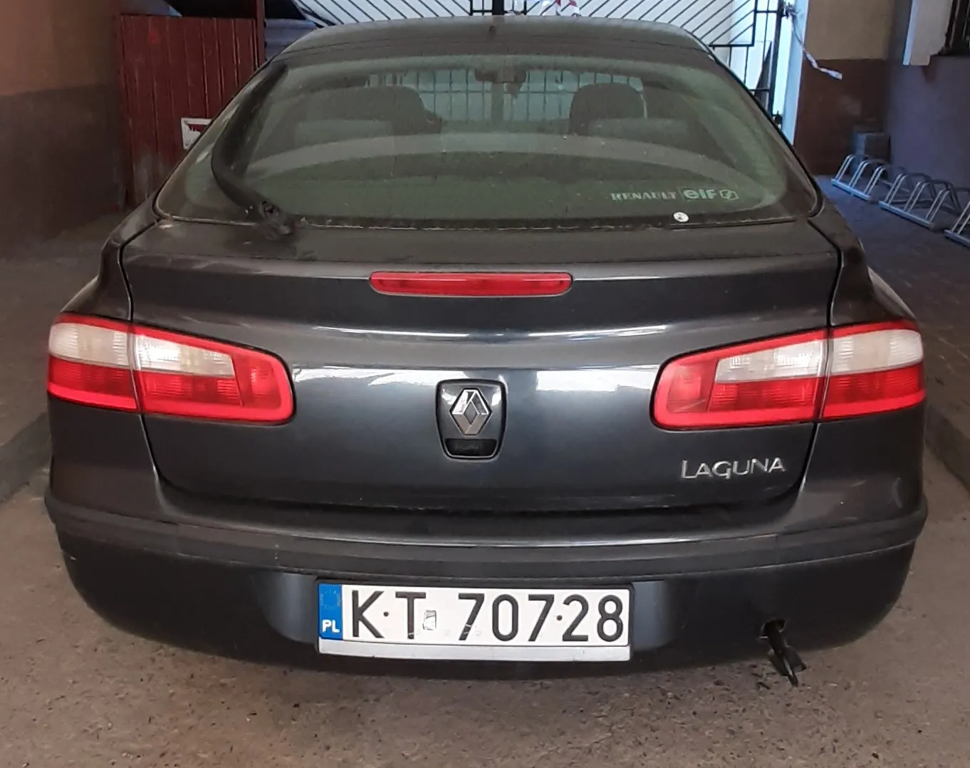 Tył ciemnoszarego samochodu marki Renault Laguna zaparkowanego na betonowej powierzchni. Widoczna tablica rejestracyjna z polskim kodem PL i numerem KT 70728. Na bagażniku napis LAGUNA oraz logo Renault. Czerwone światła tylnego oświetlenia, czerwony pasek świetlny na szybie. W tle czerwone drzwi, metalowe stojaki na rowery i fragment budynku z kratą.