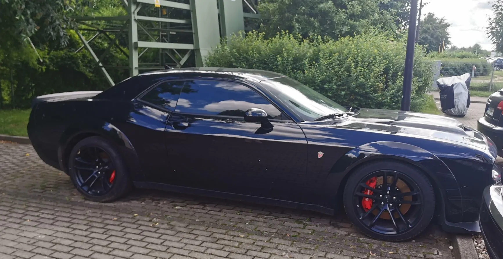 Czarny samochód sportowy typu muscle car zaparkowany na kostce brukowej. Pojazd ma czarne felgi z widocznymi czerwonymi zaciskami hamulcowymi. W tle widać zielone krzewy, drzewa oraz fragment zielonej konstrukcji stalowej.