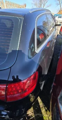 Czarny samochód widziany od tyłu z czerwonymi światłami tylnymi, zaparkowany na trawie. Obok znajduje się czerwony samochód. W tle drzewa i budynek z ogrodzeniem. Jasne niebo.