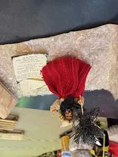 Lalka w czerwonej sukience i czarnych włosach leżąca na teksturowanej powierzchni. W tle widoczne są stare dokumenty lub książki, żółty przedmiot oraz różne przedmioty.