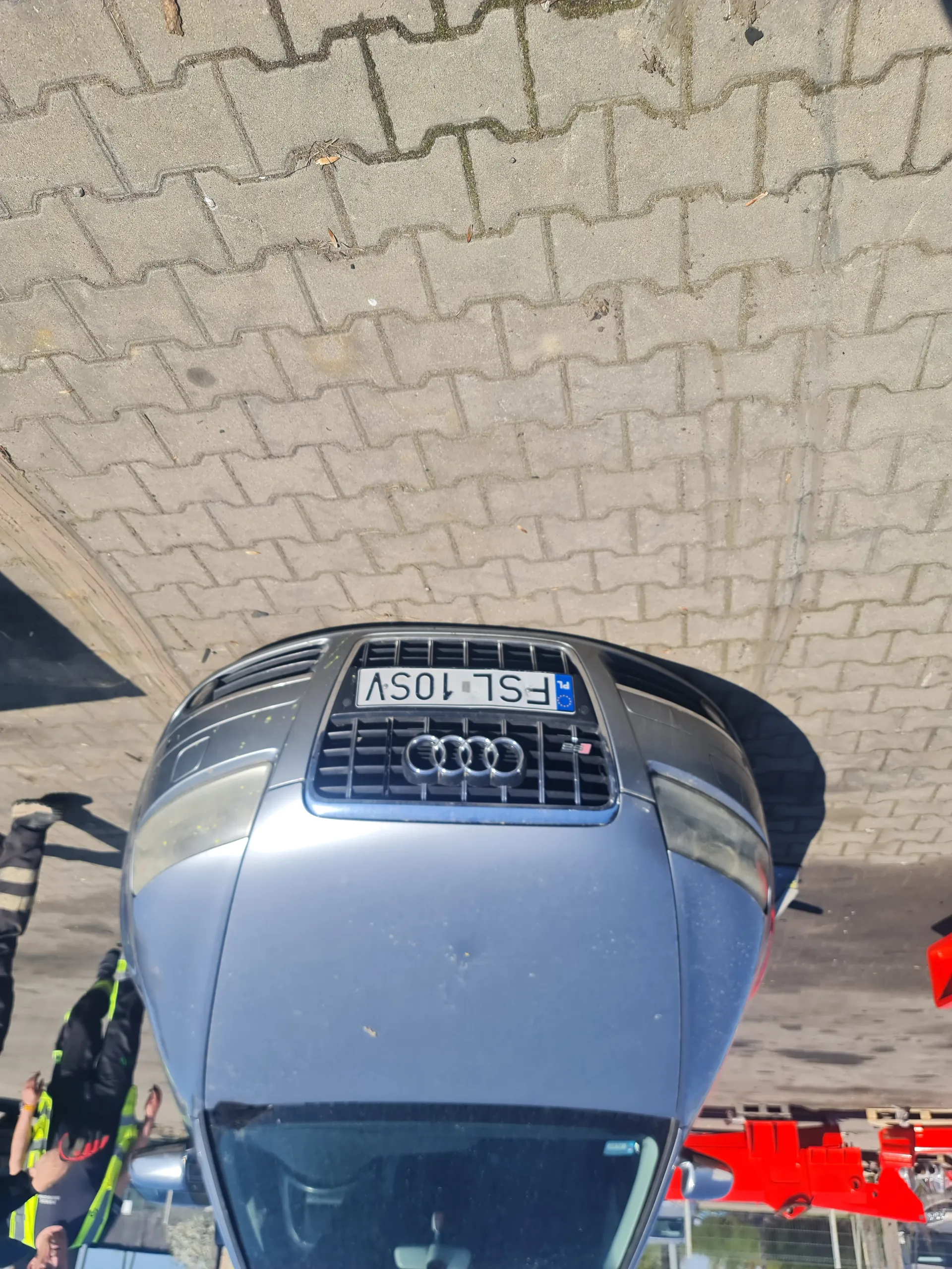 Przednia część szarego samochodu marki Audi z widocznym logo i tablicą rejestracyjną FSL 10SV. W tle osoby w żółto-zielonych kamizelkach odblaskowych oraz brukowana powierzchnia. Po lewej stronie widoczny czerwony obiekt.