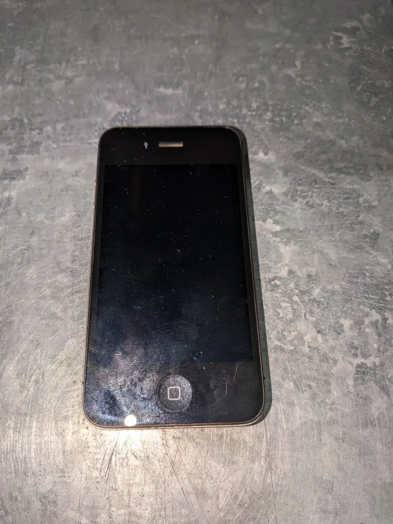Czarny smartfon z wyłączonym ekranem, leżący pionowo na szarej, porysowanej powierzchni metalowej lub kamiennej. Urządzenie posiada okrągły przycisk główny na dole ekranu.