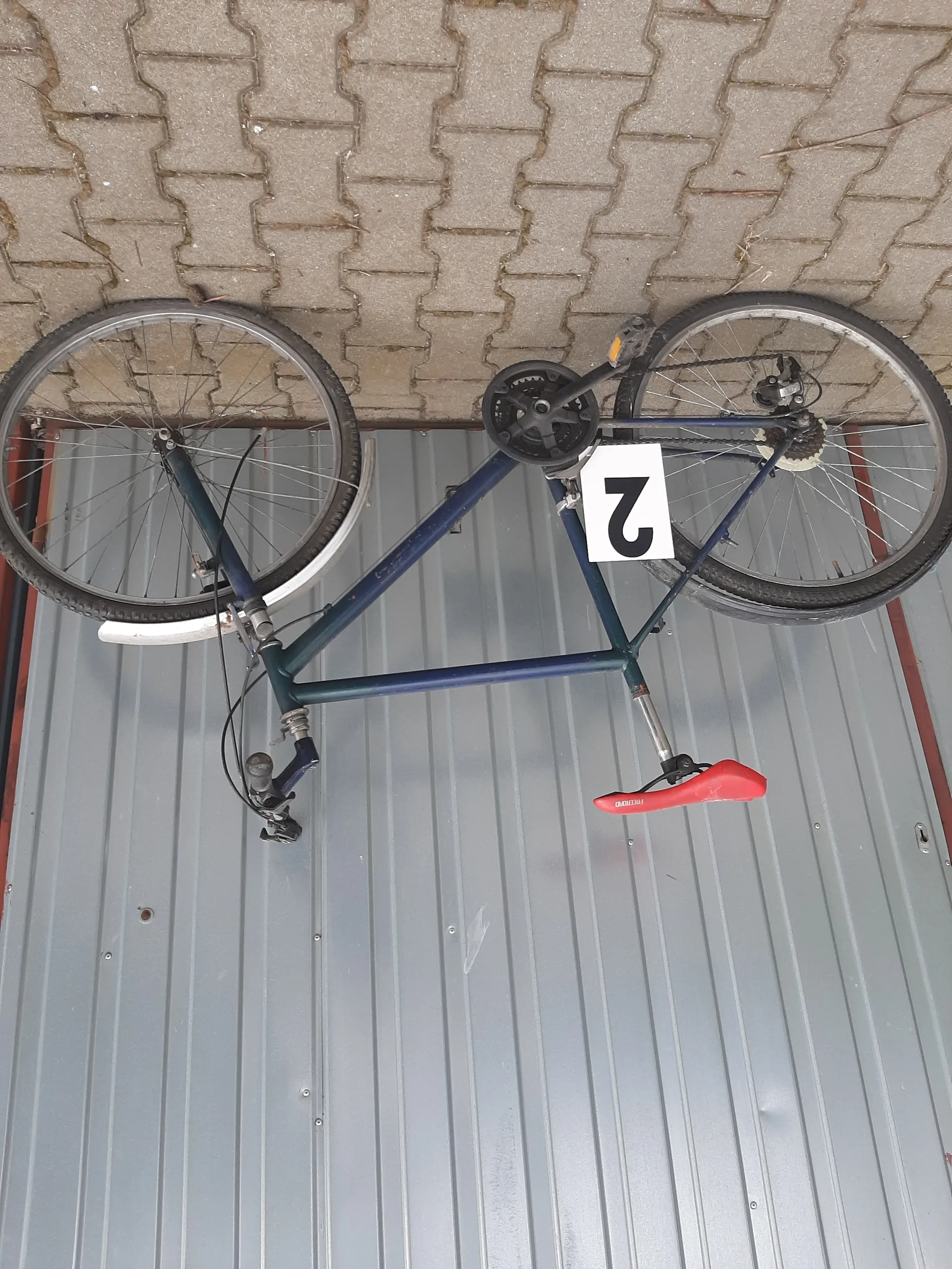Niebieski rower z czerwonym siedziskiem oparty o szarą metalową bramę garażową i brukowaną nawierzchnię. Na ramie roweru umieszczono białą tabliczkę z czarną cyfrą 2. Widoczne są koła, kierownica oraz mechanizm przerzutki.
