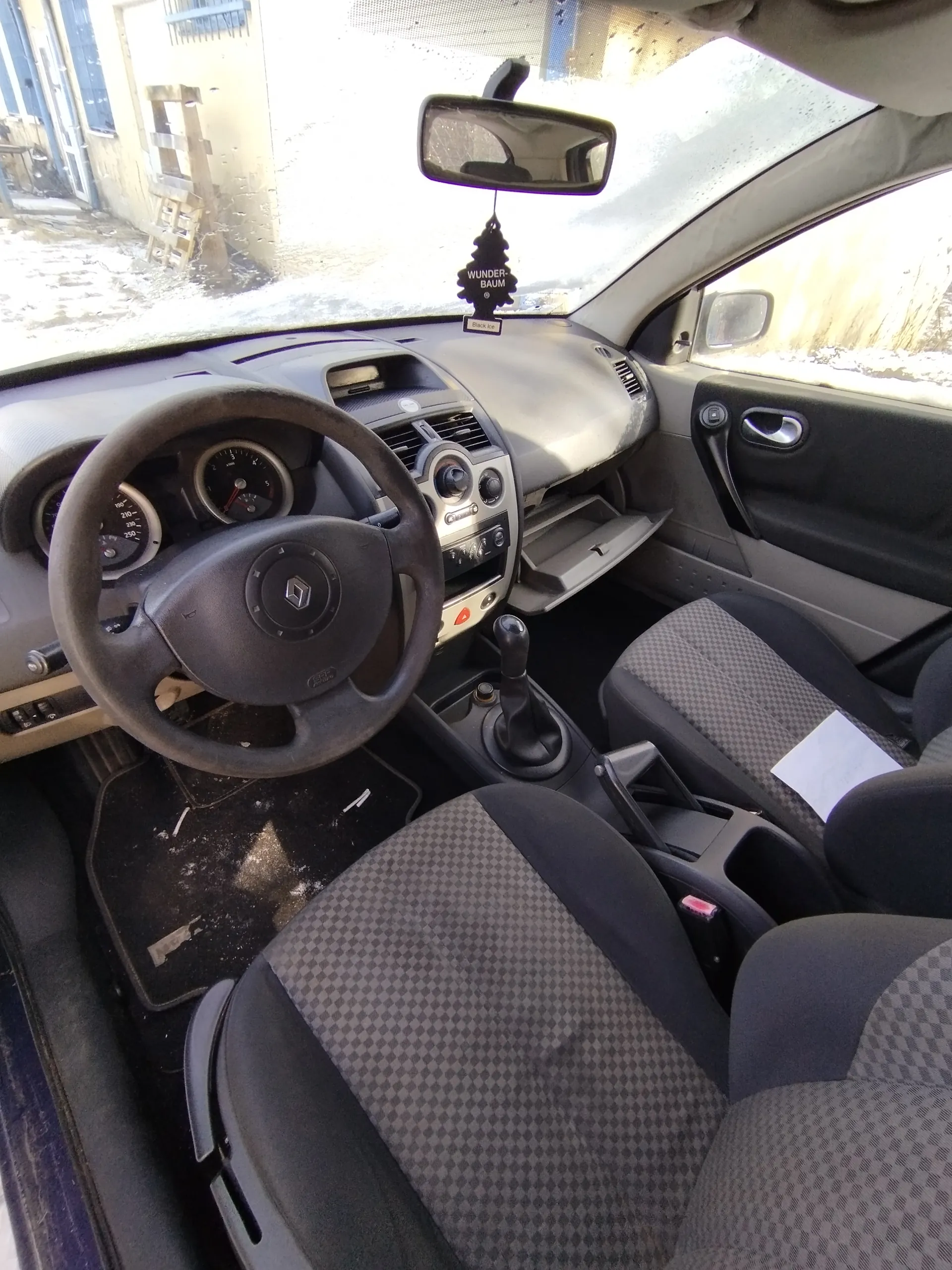 Wnętrze samochodu marki Renault z widocznym kierownicą, tablicą rozdzielczą, dźwignią zmiany biegów i siedzeniami w szaro-czarną kratę. Na lusterku wstecznym wiszący odświeżacz powietrza z napisem "WUNDER-BAUM". Przez przednią szybę widoczny zewnętrzny widok z budynkiem, schodami i śniegiem na ziemi. Drzwi pasażerskie są otwarte, a wewnątrz samochodu leżą różne przedmioty na podłodze.