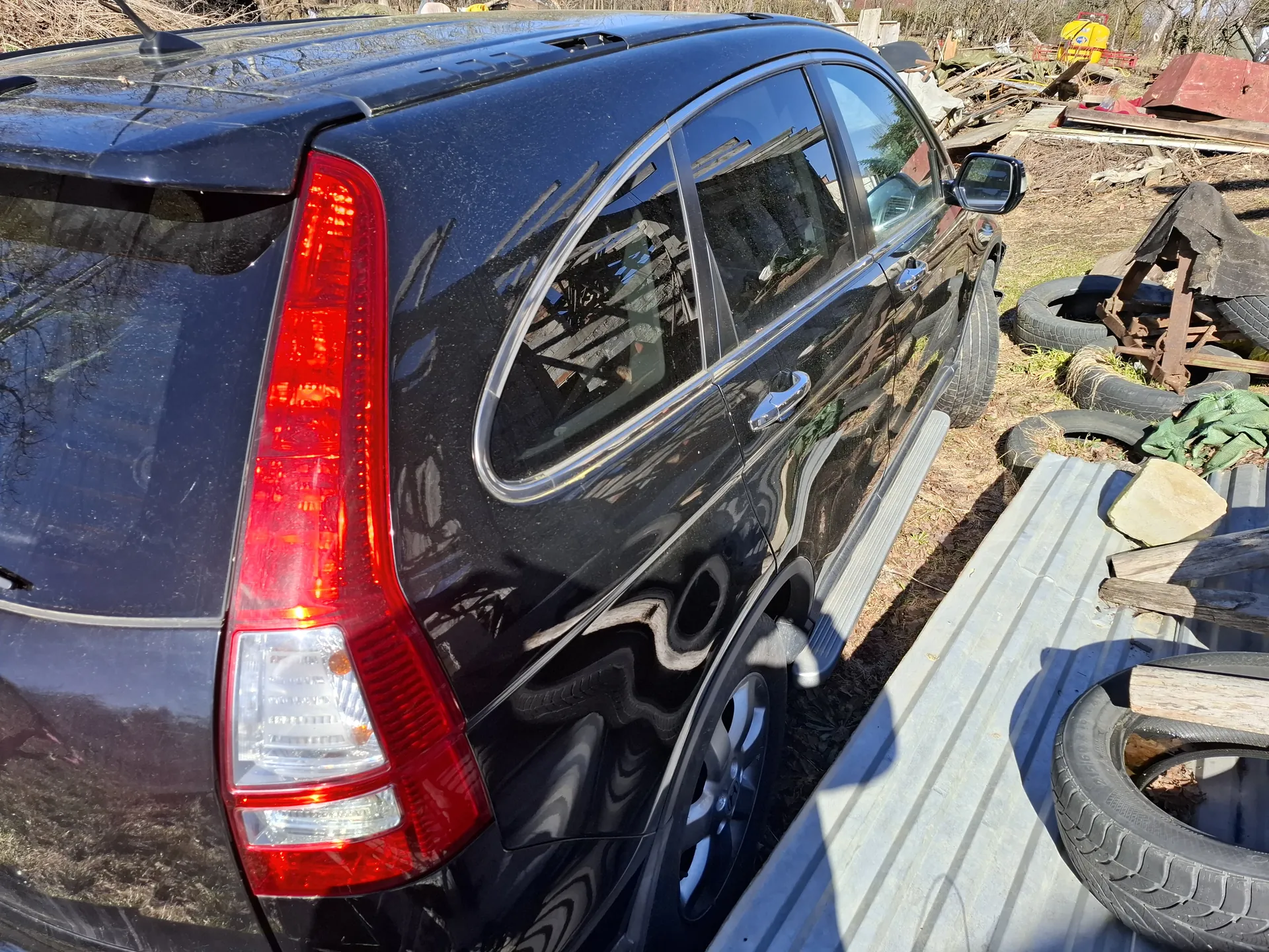 Czarny samochód typu SUV widoczny z tyłu i boku, z czerwonym światłem tylnym. Wokół pojazdu znajdują się sterty opon, drewniane deski, kamień i inne przedmioty na suchym, trawiastym terenie. W tle widoczne są suche krzewy i fragmenty budynków. Brak czytelnego tekstu.