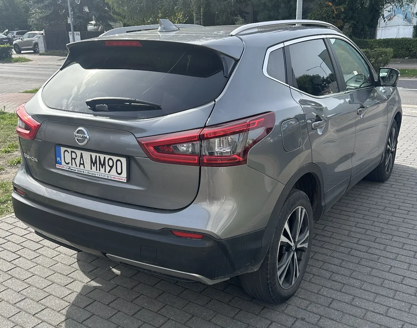 Srebrnoszary samochód marki Nissan Qashqai zaparkowany na kostce brukowej. Widok z tyłu i z boku pojazdu. Samochód posiada polską tablicę rejestracyjną o numerze CRA MM90.