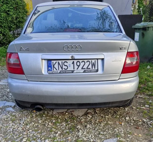 Tylny widok srebrnego samochodu marki Audi A4 1.6. Pojazd posiada polską tablicę rejestracyjną o numerze KNS 1922W. Na ramce tablicy widnieje napis 'KOBYŁKA AUTO SERWIS AUTO CZĘŚCI'. Samochód zaparkowany jest na żwirowym podłożu.