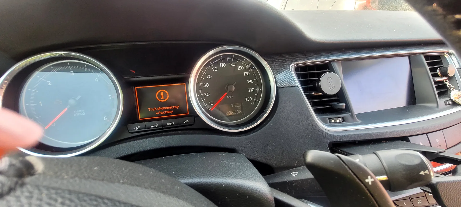 Wnętrze samochodu z widocznym kokpitem. Po lewej stronie zegar obrotomierza z czerwoną igłą, po prawej zegar prędkościomierza z czerwoną igłą. W centrum wyświetlacz z pomarańczowym komunikatem: "Tryb ekonomiczny włączony" oraz ikoną informacyjną. Poniżej komunikatu przyciski oznaczone "CHECK" i "000". Po prawej stronie konsola środkowa z ekranem, wydmuchami powietrza i kluczykiem z breloczkiem. Widoczna również część kierownicy i dźwigni sterowania.