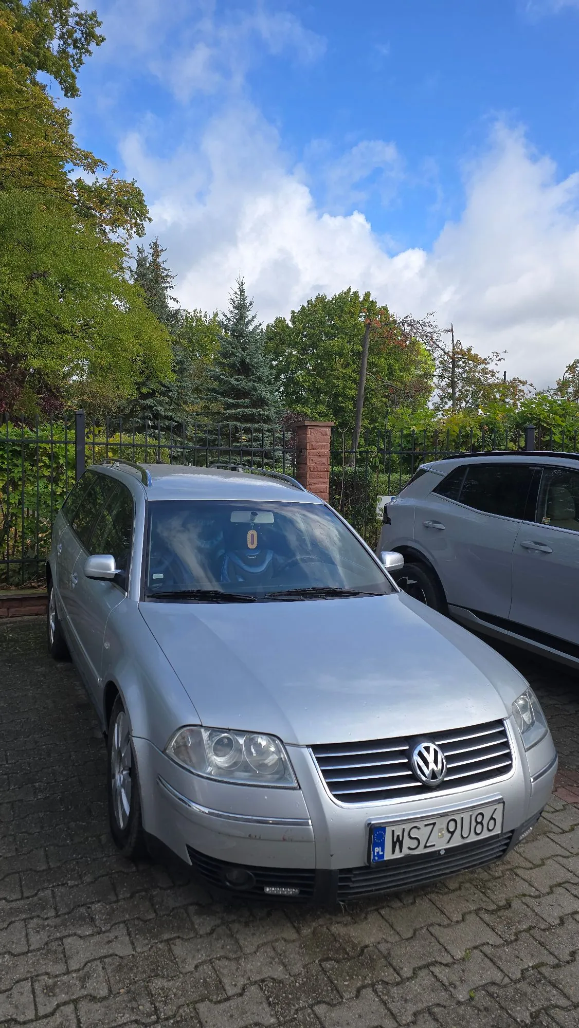 Srebrny samochód marki Volkswagen w nadwoziu kombi zaparkowany na kostce brukowej. Pojazd ma polską tablicę rejestracyjną o numerze WSZ 9U86. W tle widoczne są zielone drzewa, krzewy oraz fragment innego, jasnego samochodu po prawej stronie. Nad wszystkim znajduje się niebieskie niebo z białymi chmurami.
