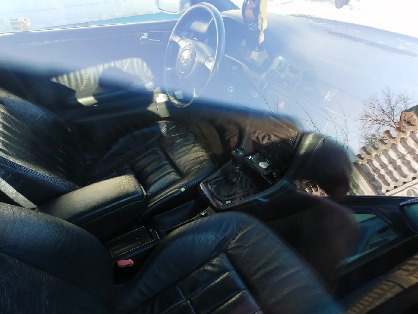 Wnętrze samochodu marki Audi z czarnymi skórzany siedzeniami, kierownicą z logo Audi, dźwignią zmiany biegów i deską rozdzielczą. Widok z pasażerskiego miejsca na stronę kierowcy. Przez przednią szybę widoczny budynek i drzewa bez liści.