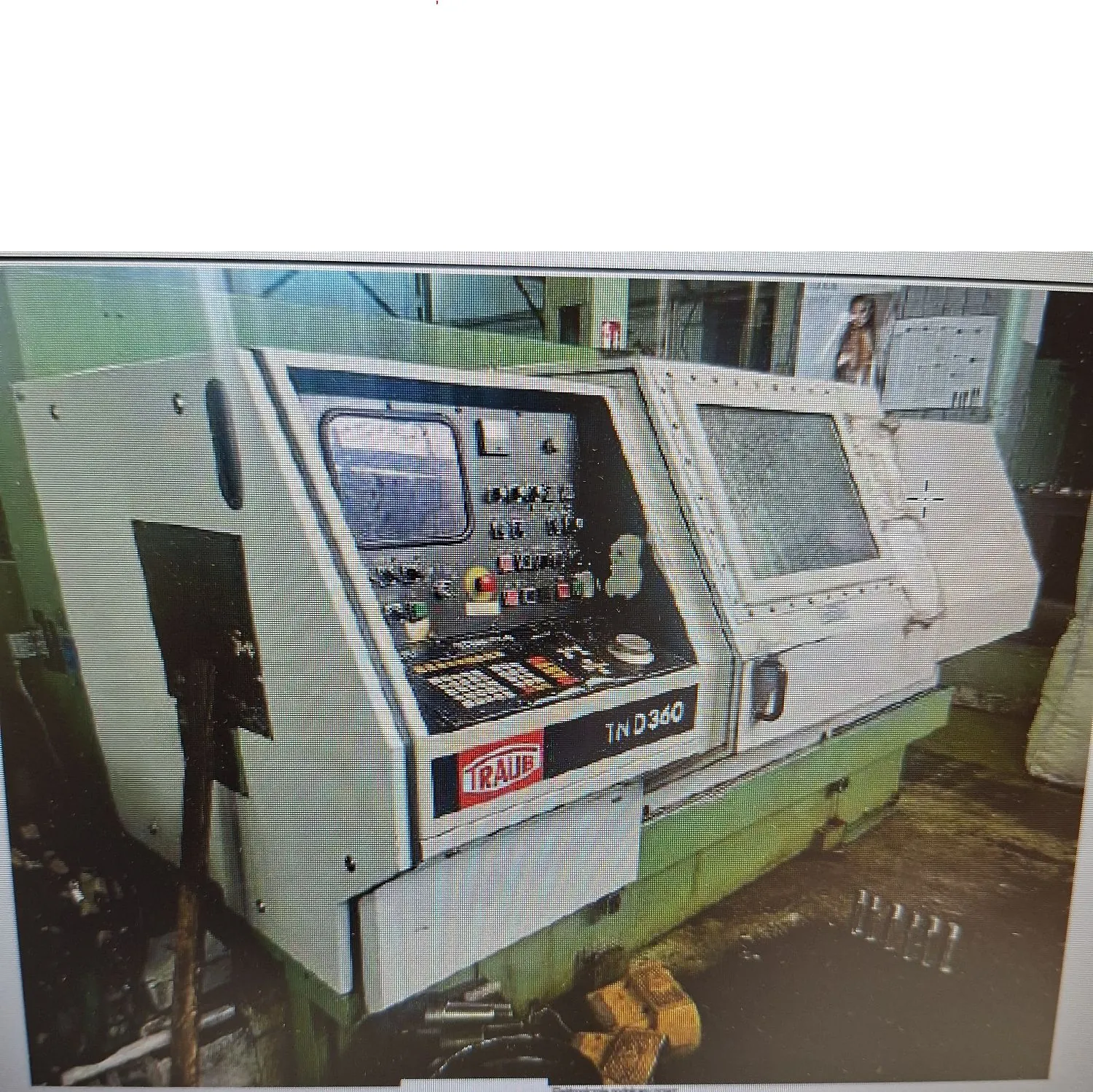 Duża, przemysłowa maszyna CNC marki Traub, model TND360. Maszyna ma jasnoszarą obudowę z panelem sterowania zawierającym ekran, liczne przyciski, przełączniki i klawiaturę. Po prawej stronie znajduje się otwarta klapa z szybą. Maszyna spoczywa na zielonej podstawie.