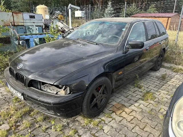 Czarny samochód marki BMW w nadwoziu kombi, zaparkowany na kostce brukowej. Pojazd jest w złym stanie technicznym: brakuje przedniego reflektora po lewej stronie, a lakier na masce jest zmatowiały i zabrudzony. W tle widoczne jest ogrodzenie z siatki, drzewa oraz fragmenty przemysłowych urządzeń i mały budynek z czerwonym dachem.
