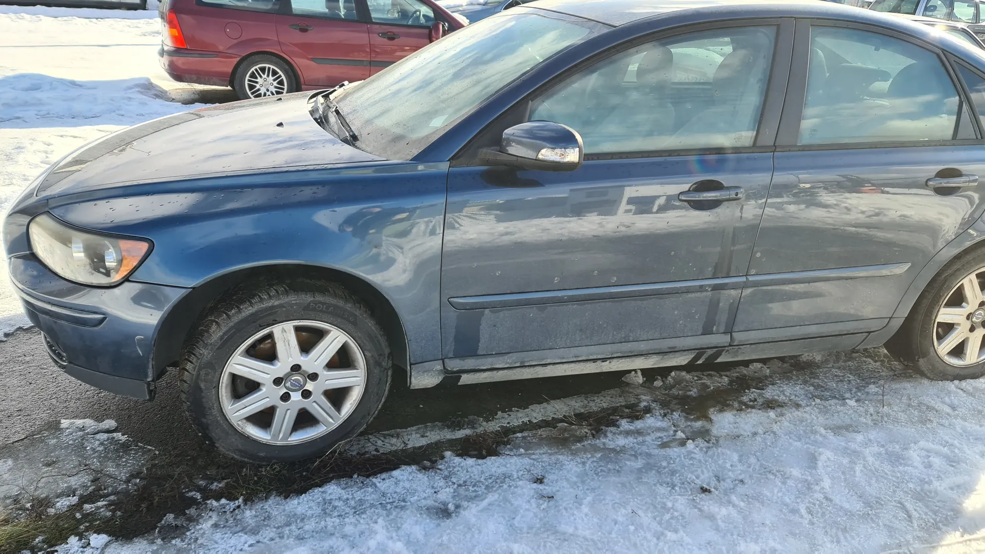 Niebieski samochód osobowy typu sedan zaparkowany na zimowym parkingu. Pojazd ma metalowe felgi, na karoserii widoczne są ślady brudu. W tle czerwony samochód, a na ziemi resztki śniegu i lodu. Tło pokazuje asfaltową powierzchnię z miejscami pokrytymi śniegiem.