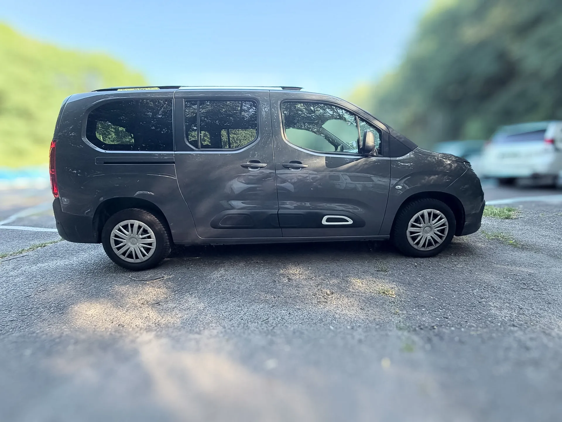 Szary samochód typu minivan zaparkowany bokiem na asfaltowym placu. Pojazd ma przesuwne drzwi boczne i srebrne felgi. W tle widoczne są rozmyte zielone drzewa oraz fragment innego białego samochodu.