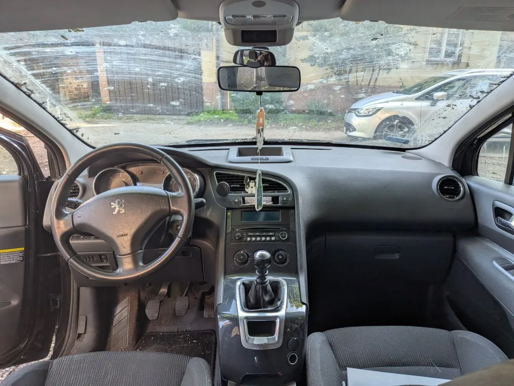 Widok z wnętrza samochodu marki Peugeot, z perspektywy kierowcy. Widoczna jest kierownica z logo lwa, deska rozdzielcza, konsola środkowa z drążkiem zmiany biegów oraz przednie fotele z ciemnej tkaniny. Przez przednią szybę, która jest mocno rozbita i popękana, widać zaparkowany srebrny samochód oraz fragmenty otoczenia. Do lusterka wstecznego zawieszona jest ozdoba.