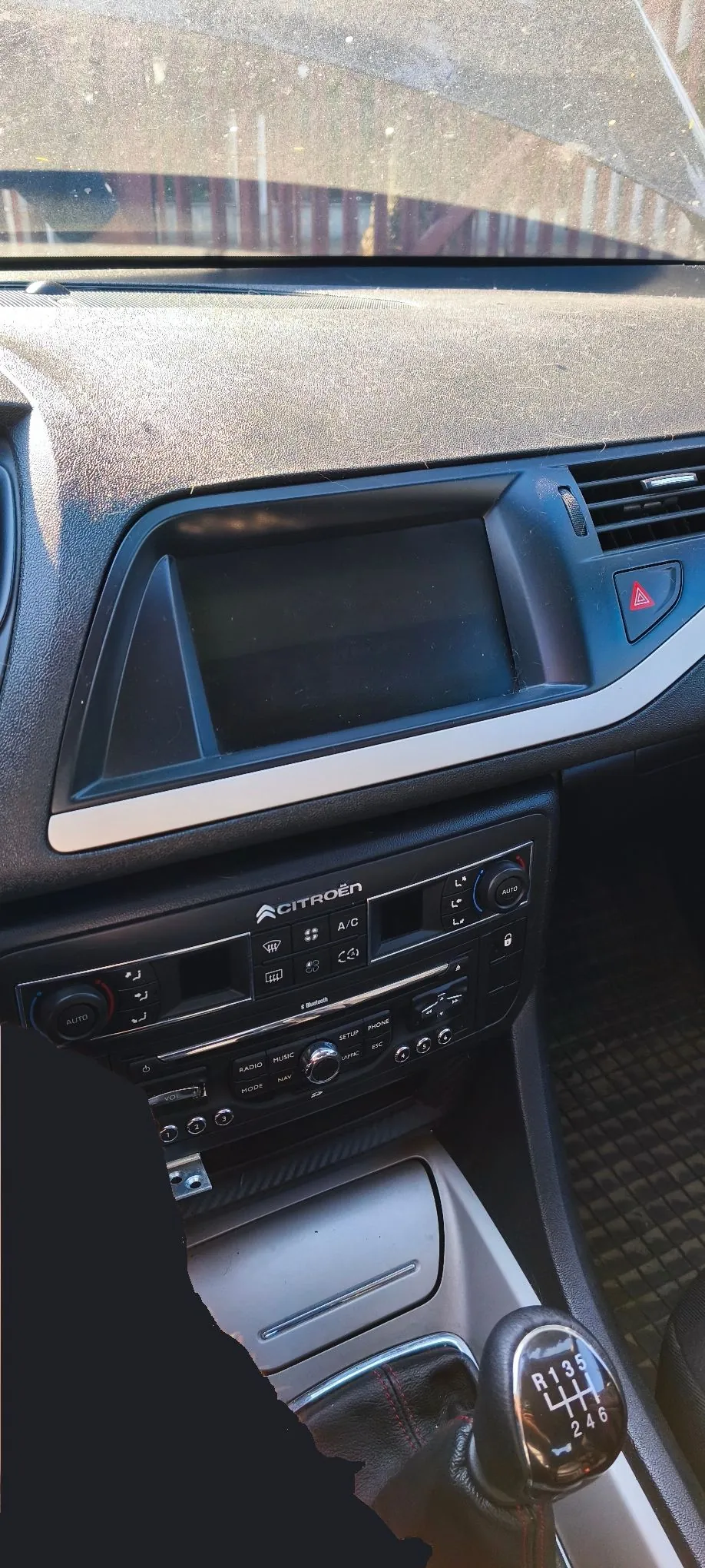 Wnętrze samochodu marki Citroën, widok na deskę rozdzielczą. W centralnej części znajduje się wyłączony ekran multimedialny w czarnej ramce z srebrnym wykończeniem u dołu. Poniżej znajduje się panel sterowania klimatyzacją z przyciskami takimi jak 'AUTO', 'A/C' oraz pokrętłami. Jeszcze niżej znajduje się radio z przyciskami 'RADIO', 'MUSIC', 'PHONE', 'NAV'. Na dole widoczna jest lewarek zmiany biegów z oznaczeniami biegów R, 1, 3, 5, 2, 4, 6.