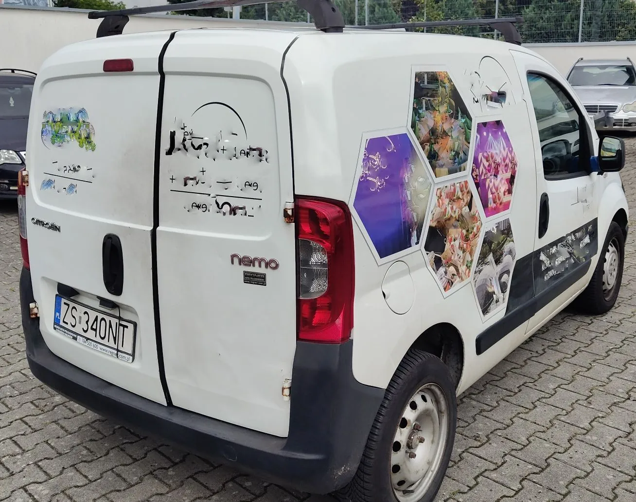 Biały samochód dostawczy marki Citroën Nemo zaparkowany na kostce brukowej. Na tylnych drzwiach i boku pojazdu znajdują się kolorowe naklejki w kształcie sześciokątów z różnymi grafikami oraz czarne napisy i symbole. Samochód ma polską tablicę rejestracyjną o numerze ZS 340NT.